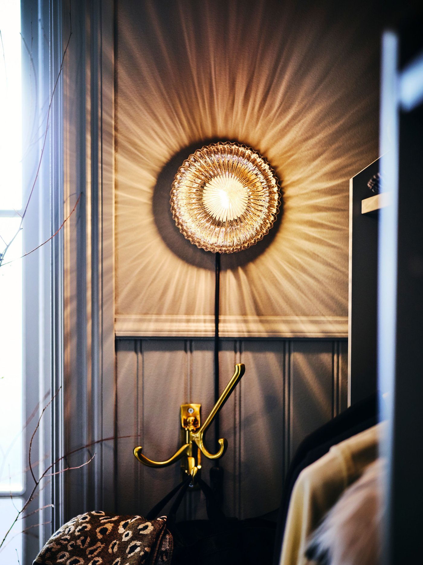 A SOLKLINT wall lamp is mounted on a wall above a KÄMPIG 3-arm hook. The lamp makes patterns of light and shadow on the wall.