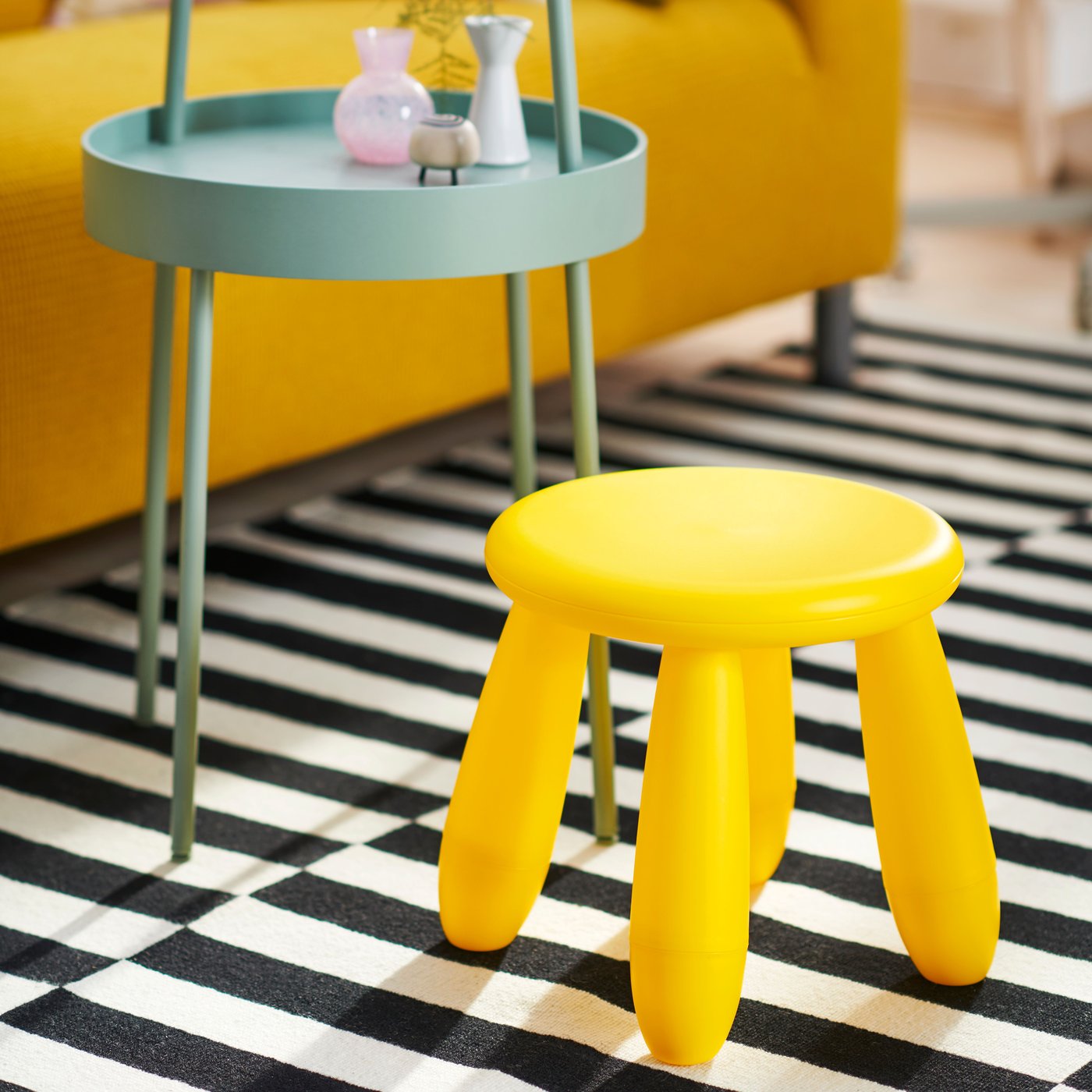 A yellow MAMMUT children’s stool and a light grey-green BURVIK side table on top of a black/white stripe STOCKHOLM rug.