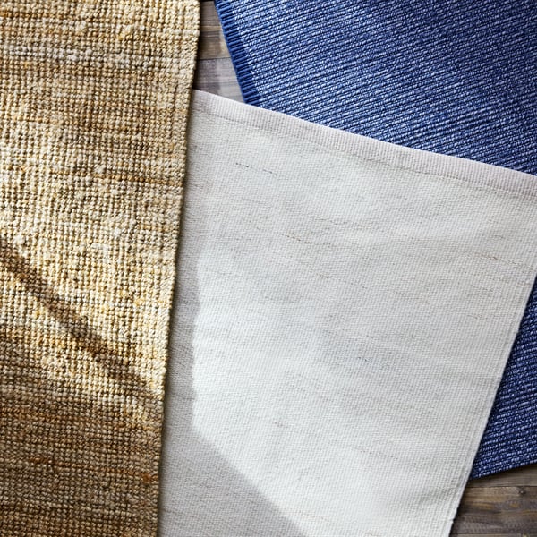 A beige flatwoven rug with a textured surface lies on top of a white rug and a blue rug on a wooden floor.