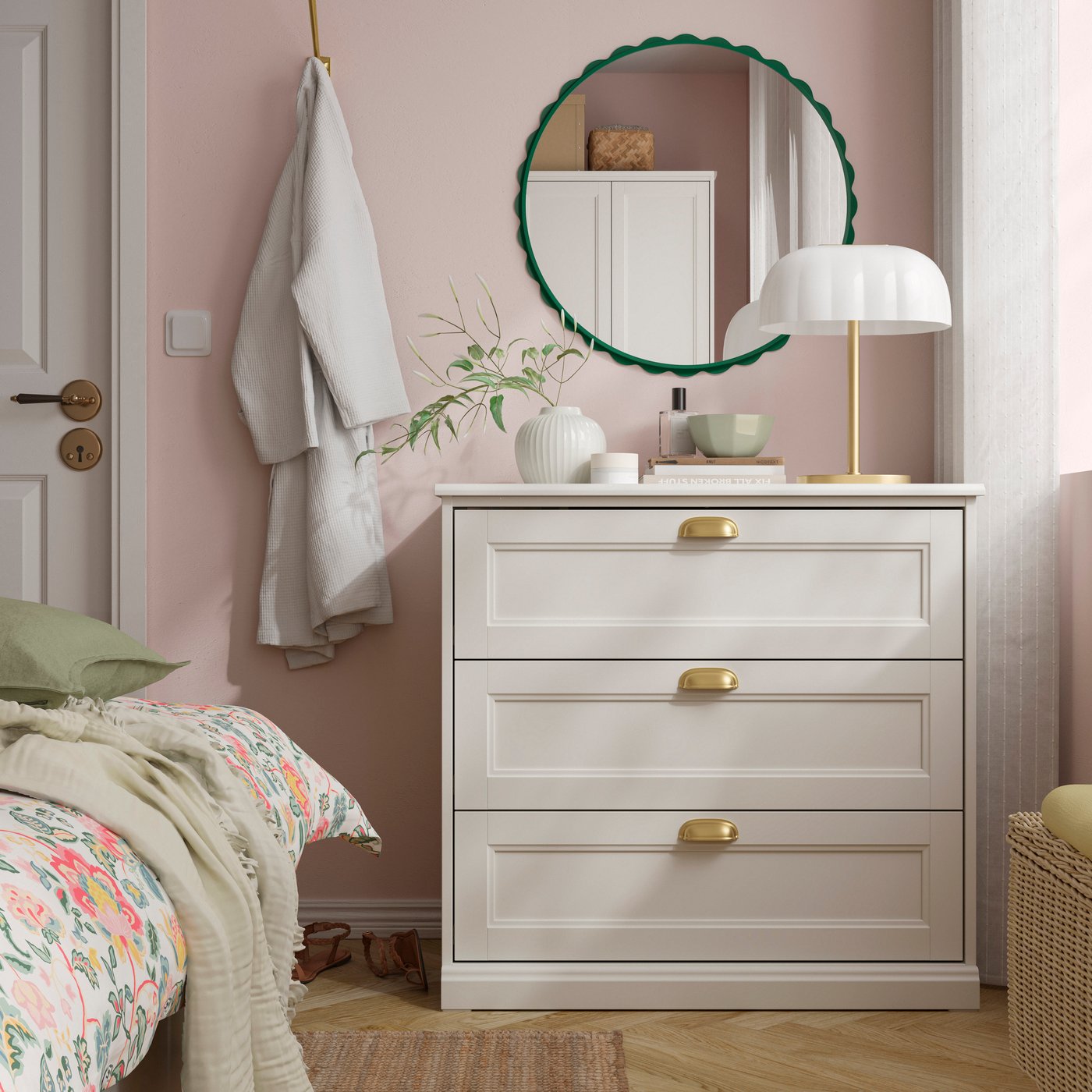 A white three-drawer dresser with gold handles sits against a blush pink wall, topped with decor and a round mirror in a cozy bedroom.