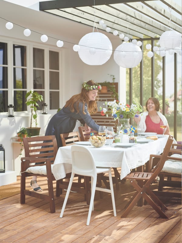 Un portico con una tavola apparecchiata con sedie e una sedia junior URBAN. Attorno, due donne che celebrano il Midsommar. Al di sopra, luci e lampade decorative.
