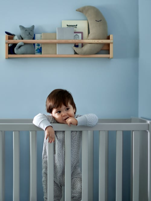 A child in a grey cot is leaning on one of the cot's sides. A wall storage with books and toys is mounted behind the child.