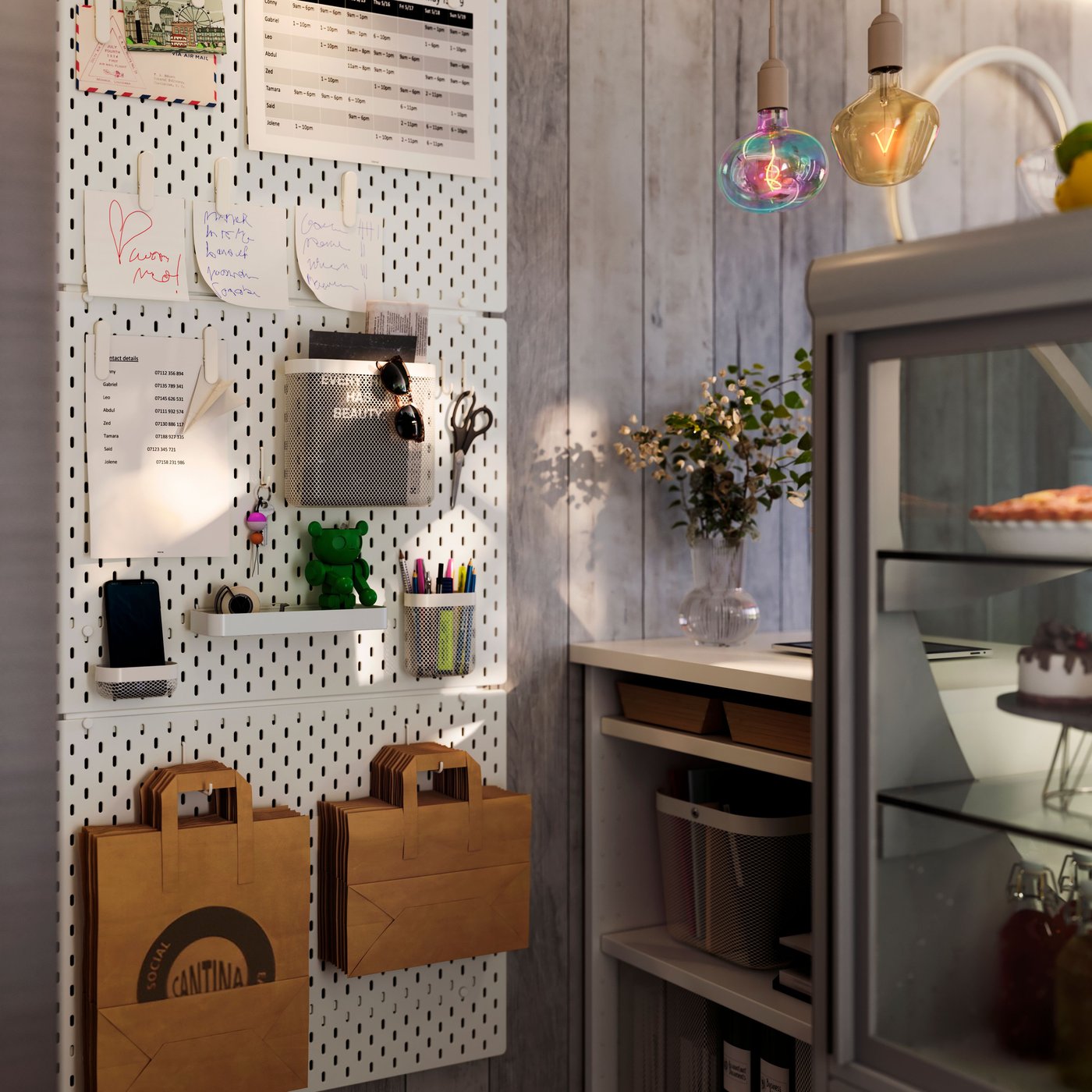 Rum med SKÅDIS anslagstavla med anteckningar och hyllor, papperspåsar under, bänk med blommor och kyl med tårtor till höger.