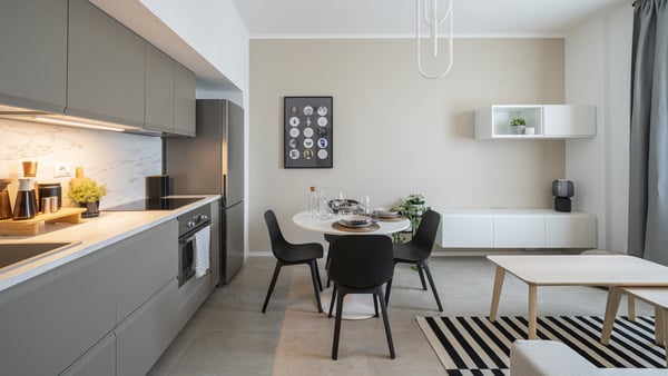 Una cucina con soggiorno annesso in cui si vede una cucina lineare e una zona con tavolo da pranzo e 4 sedie. Di lato a destra si intravede una zona soggiorno con dei tavolini in legno e un mobile TV bianco a parete.