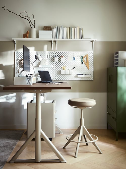 A beige sit/stand desk in a beige room with a laptop and monitor on top and a TROLLBERGET active sit/stand support in front.