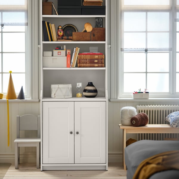 Part of a living-room where a white HAUGA high two-door cabinet holding storage and decoration stands between two windows.