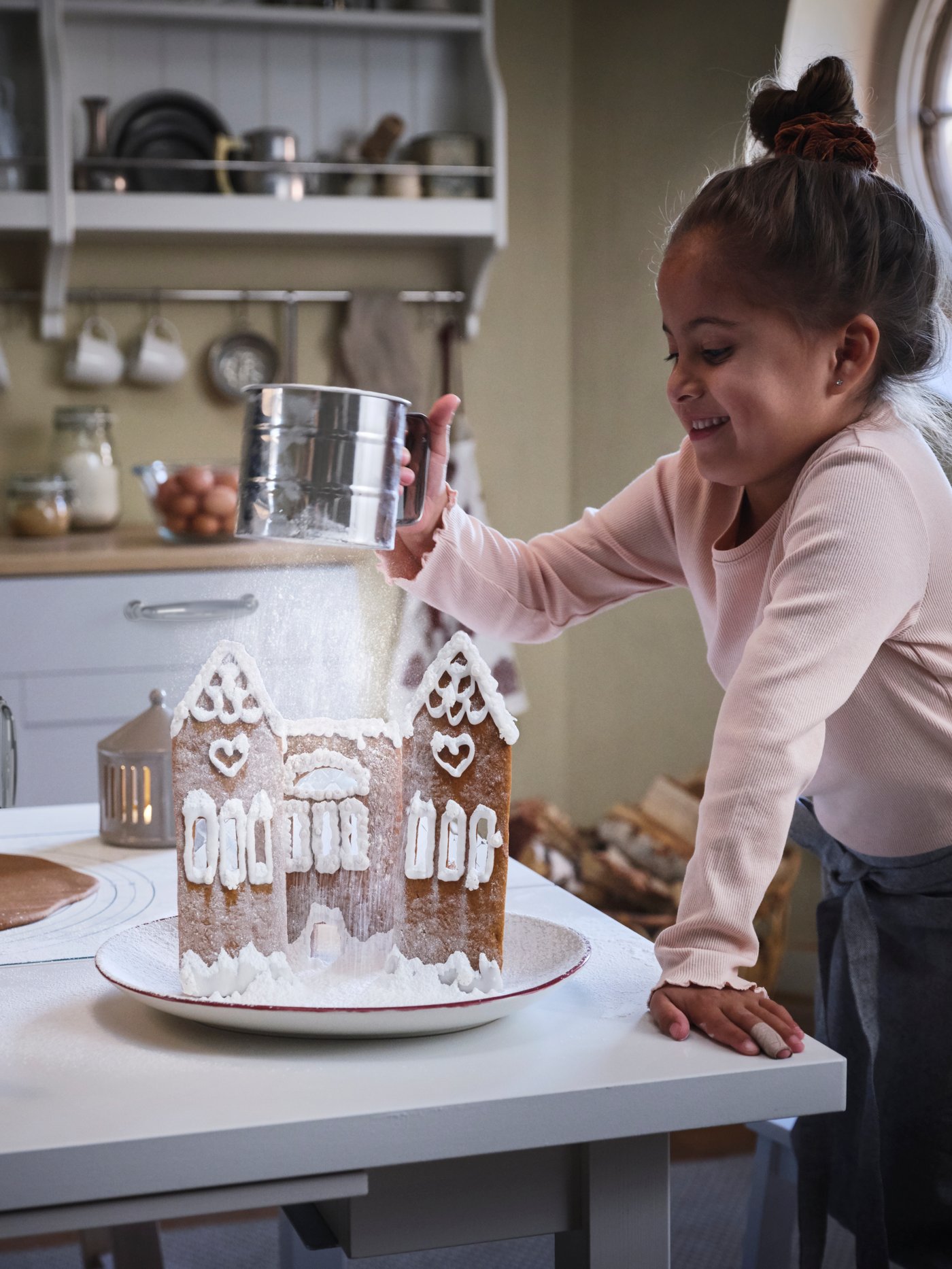 Una niña utiliza un tamizador de harina para tamizar azúcar en polvo sobre una casa de pan de jengibre.