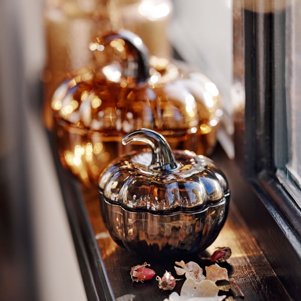 Two HÖSTAGILLE pumpkin-shaped bowls with lid, one brown and the other green-brown, are on a windowsill next to dried leaves.