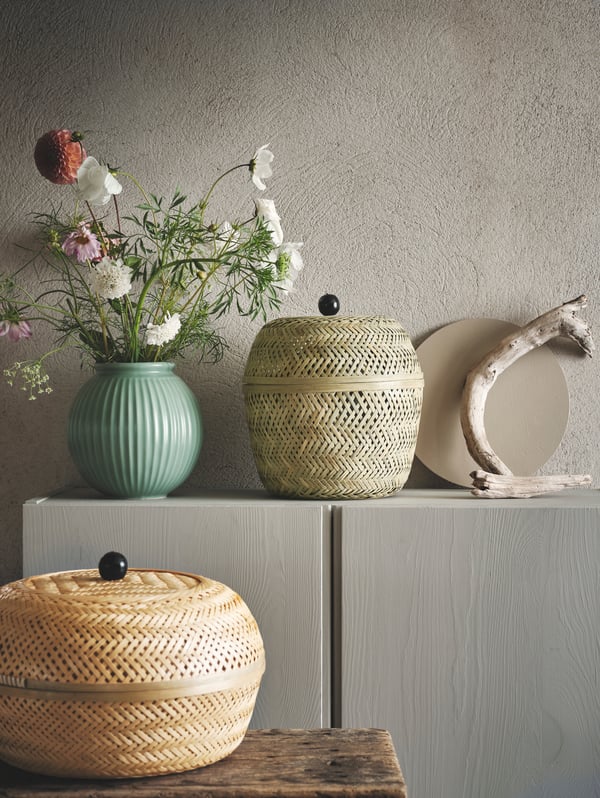 Um vaso com flores, um cesto TJILLEVIPS e uma escultura de madeira sobre um armário, por trás de um cesto TJILLEVIPS sobre uma mesa.