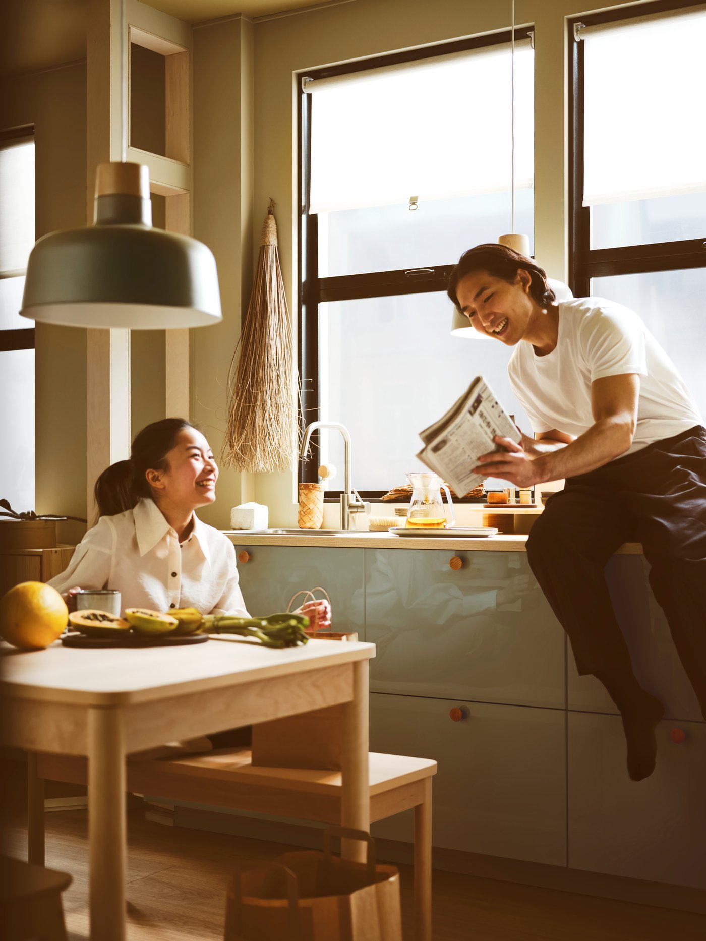 Dve osebi klepetata v kuhinji s sivo-modrimi KALLARP ličnicami, omaricami, delovno ploščo, koritom in mizo s klopjo.