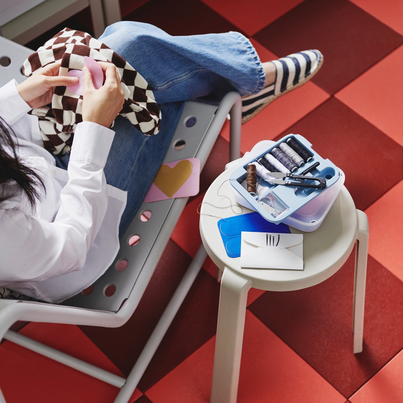 Een TRIXIG reparatieset van 39 delen ligt open op een bijzettafeltje, naast iemand in een stoel die een roze, hartvormige patch op een shirt naait.