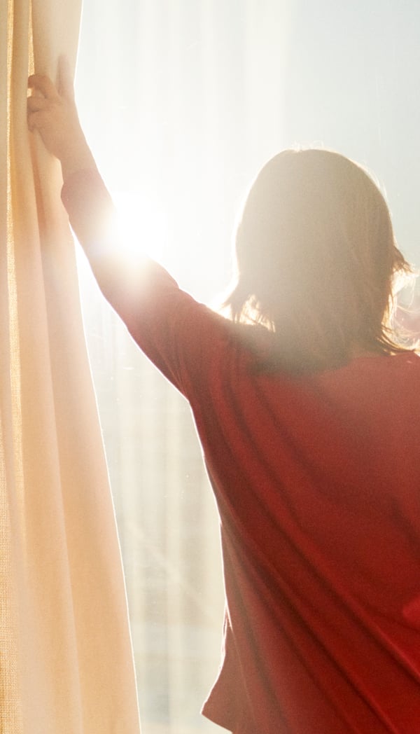 O femeie cu o cămașă roșie trage perdeaua unei ferestre inundate de lumina soarelui.