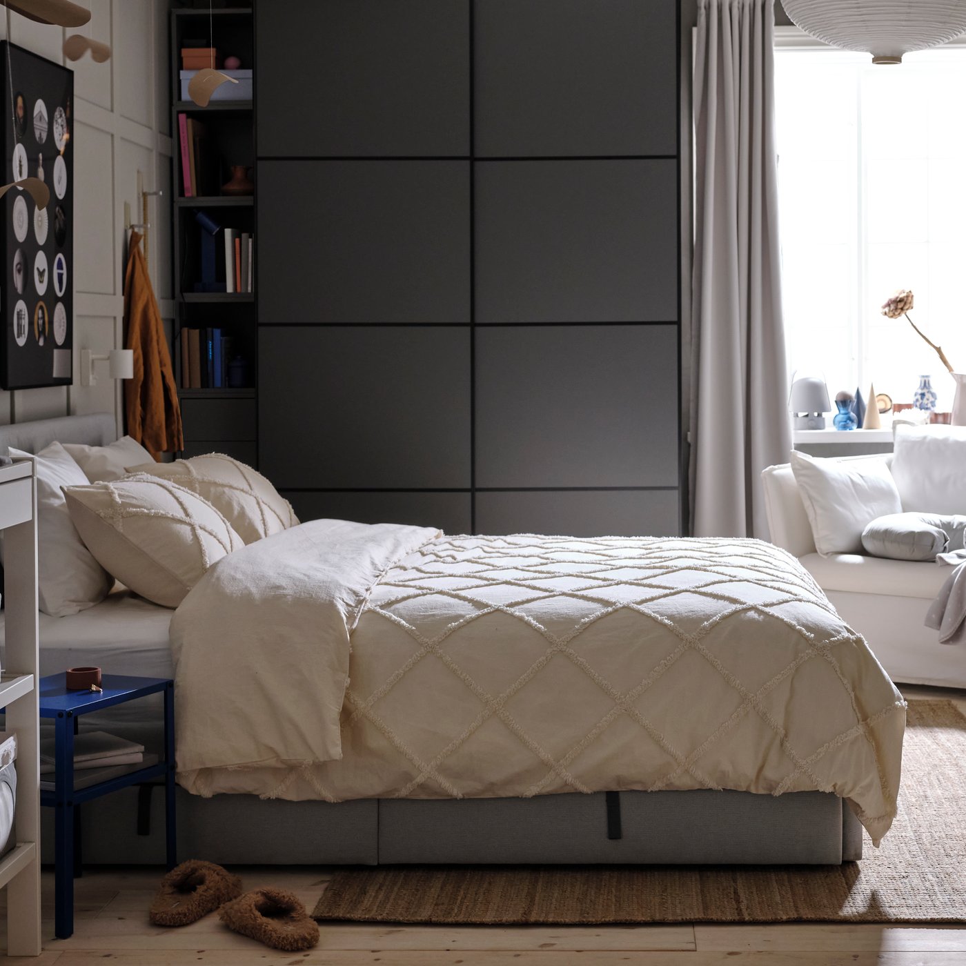 Un letto imbottito grigio, con biancheria NYPONLUGGMAL, di fronte a un guardaroba PAX/MEHAMN grigio scuro e a una poltrona bianca.