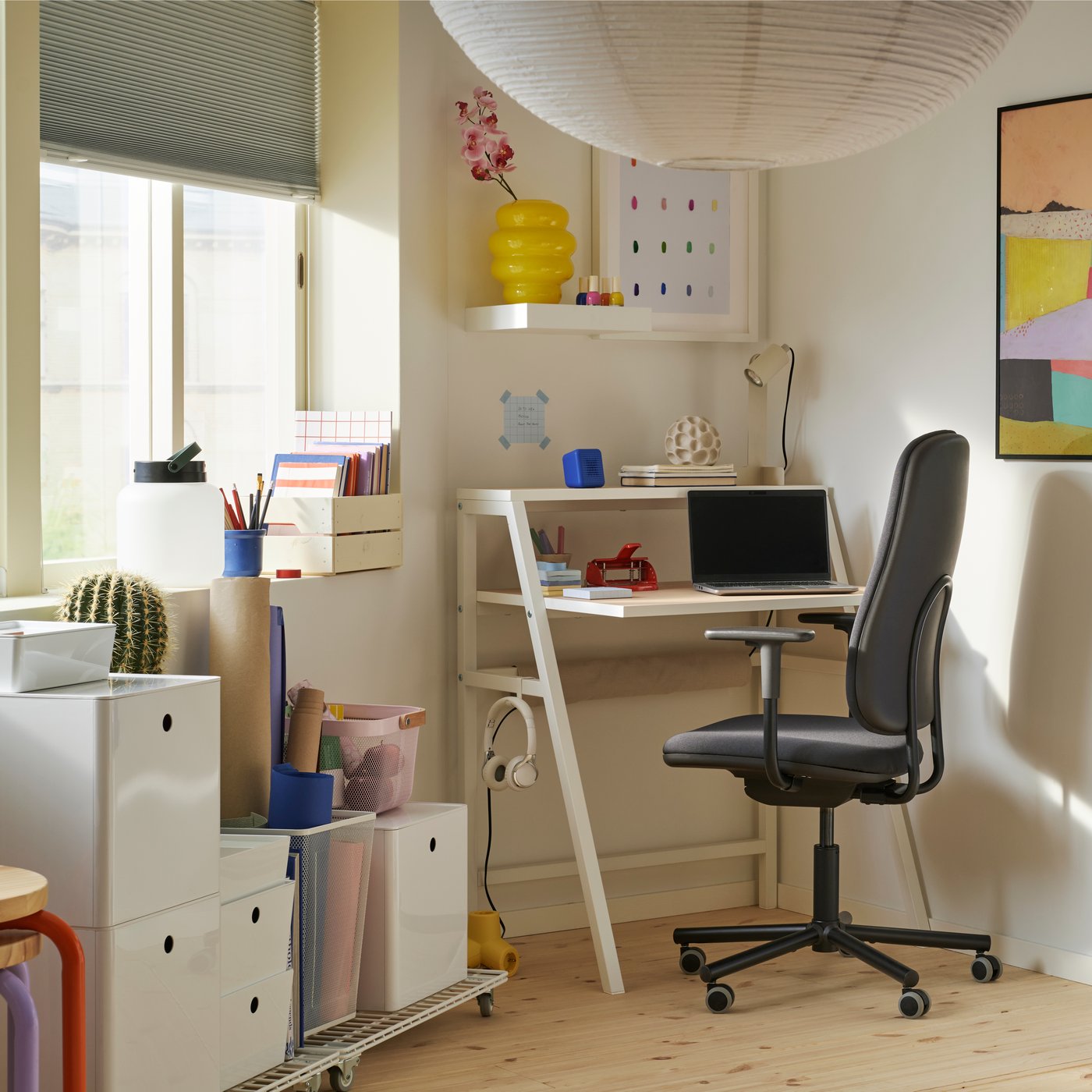 Nell'angolo di una stanza, una finestra e un piccolo spazio di lavoro con una scrivania MITTPLAN, una sedia da ufficio, uno scaffale a giorno e altre soluzioni con le rotelle per tenere tutto in ordine.