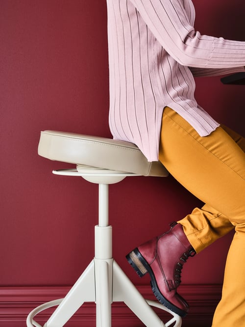 Close-up of person with pink shirt, orange pants and red boots sitting on a white stool with hands leaning on desk.