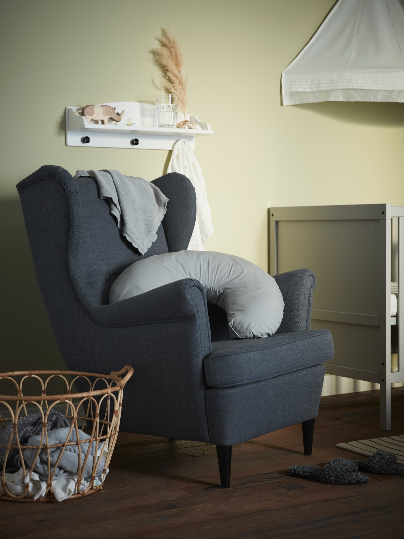 Hämärässä vauvan huoneessa on harmaa STRANDMON-nojatuoli, jonka päällä on LEN-imetystyyny.