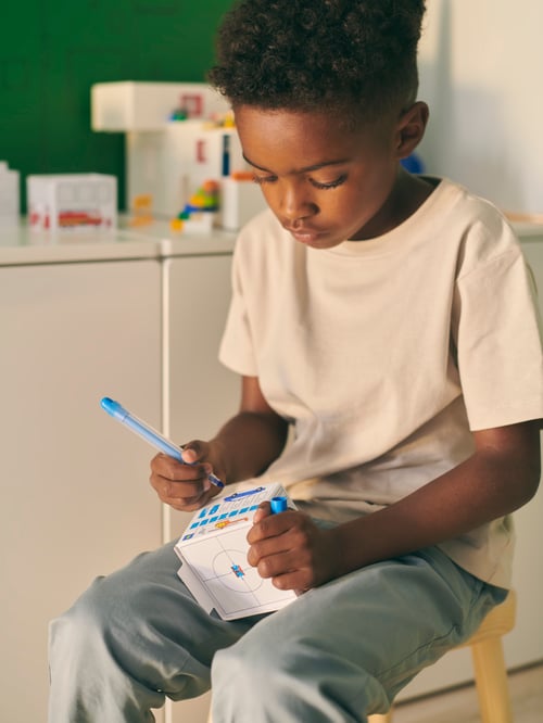 スツールに座っている男の子が、MÅLA/モーラ 厚紙でつくる街セットで折った紙の家を膝にのせて色を塗っている。