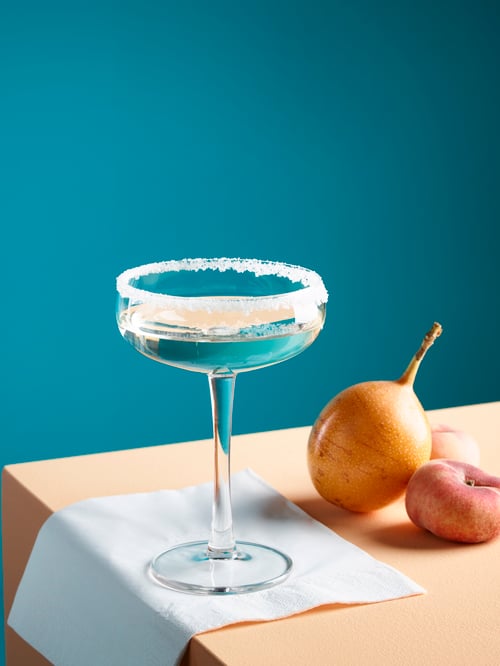 Close-up of table with champagne cup with sugared rim, on a paper napkin next to different sized fruits.