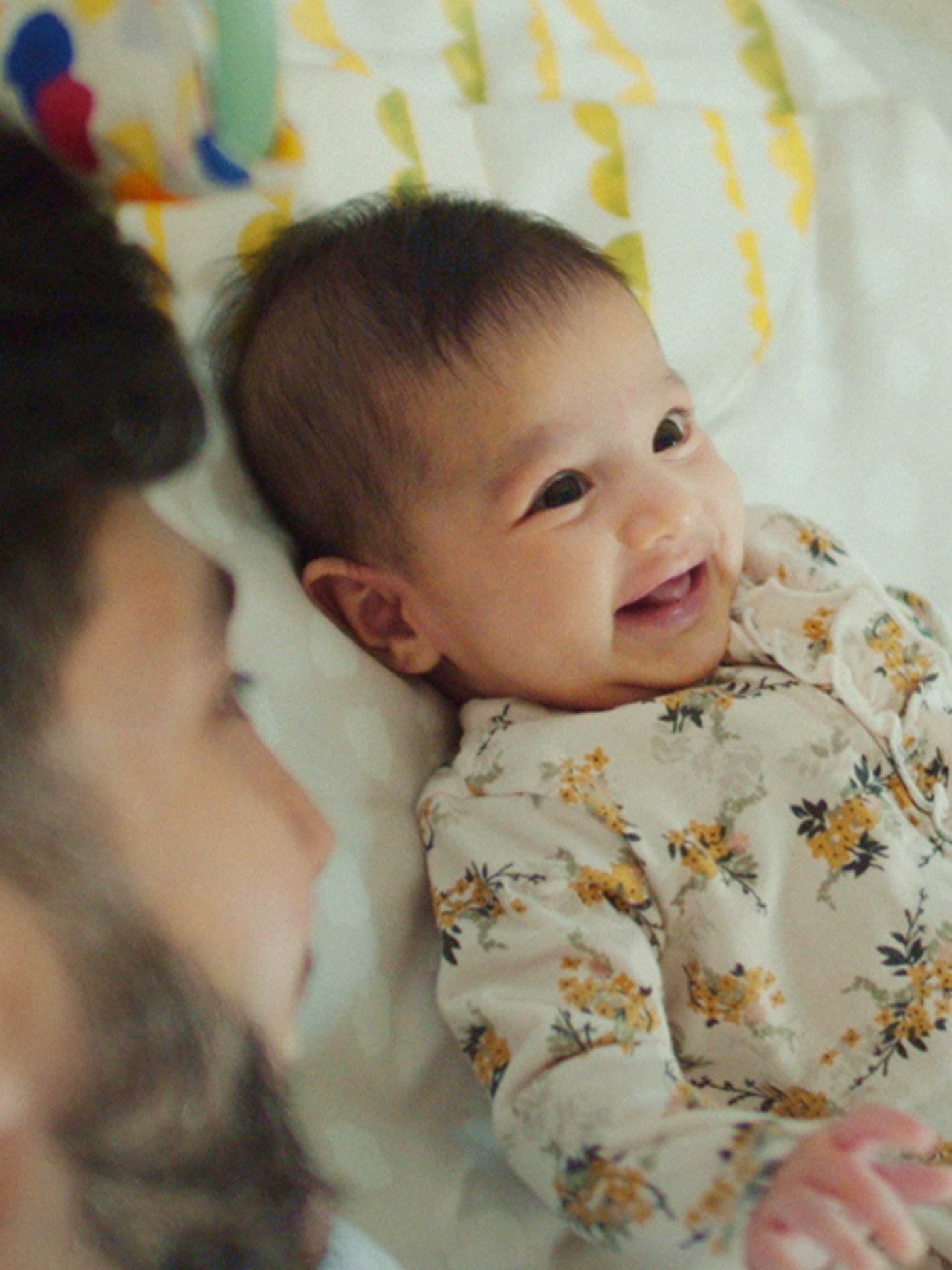 A baby with dark hair and wearing a floral jumpsuit lies on his back and smiles while his Dad looks on.