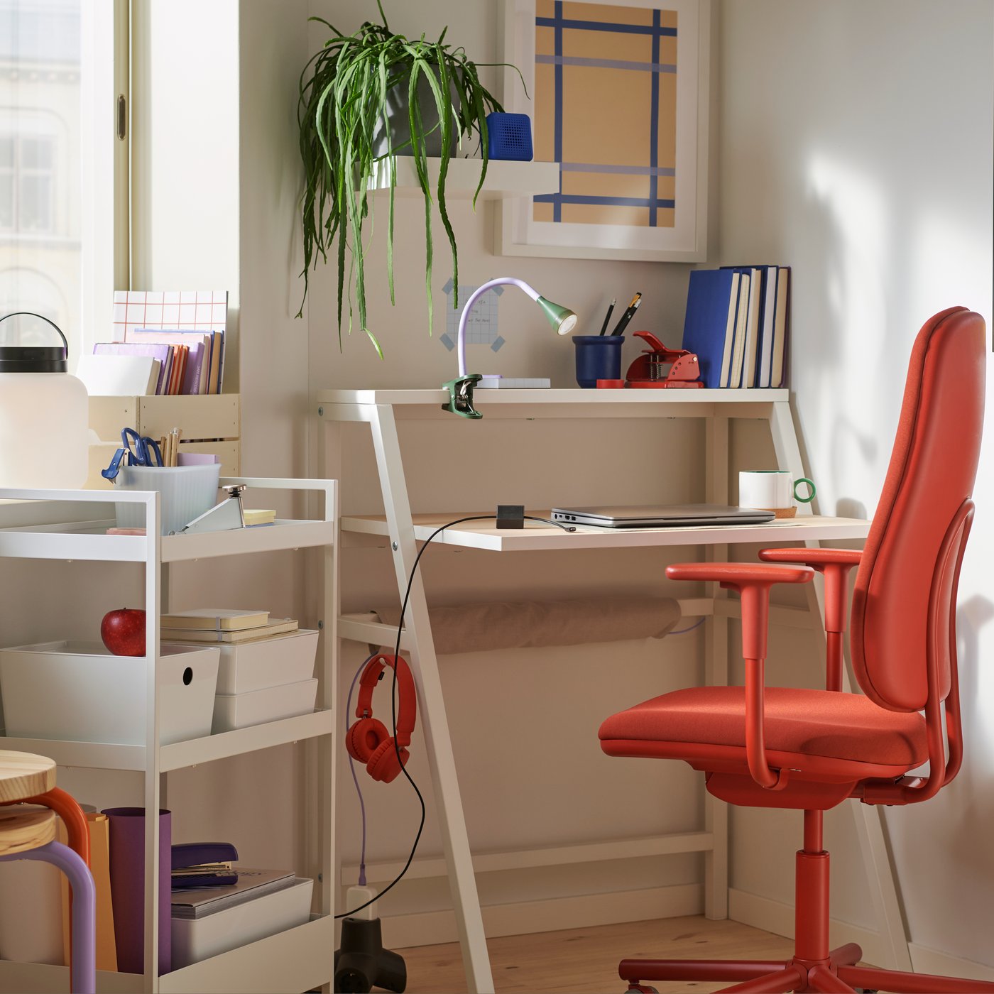 A small corner workspace with a MITTPLAN desk, a red SMÖRKULL office chair, a plant, and a white trolley with storage box