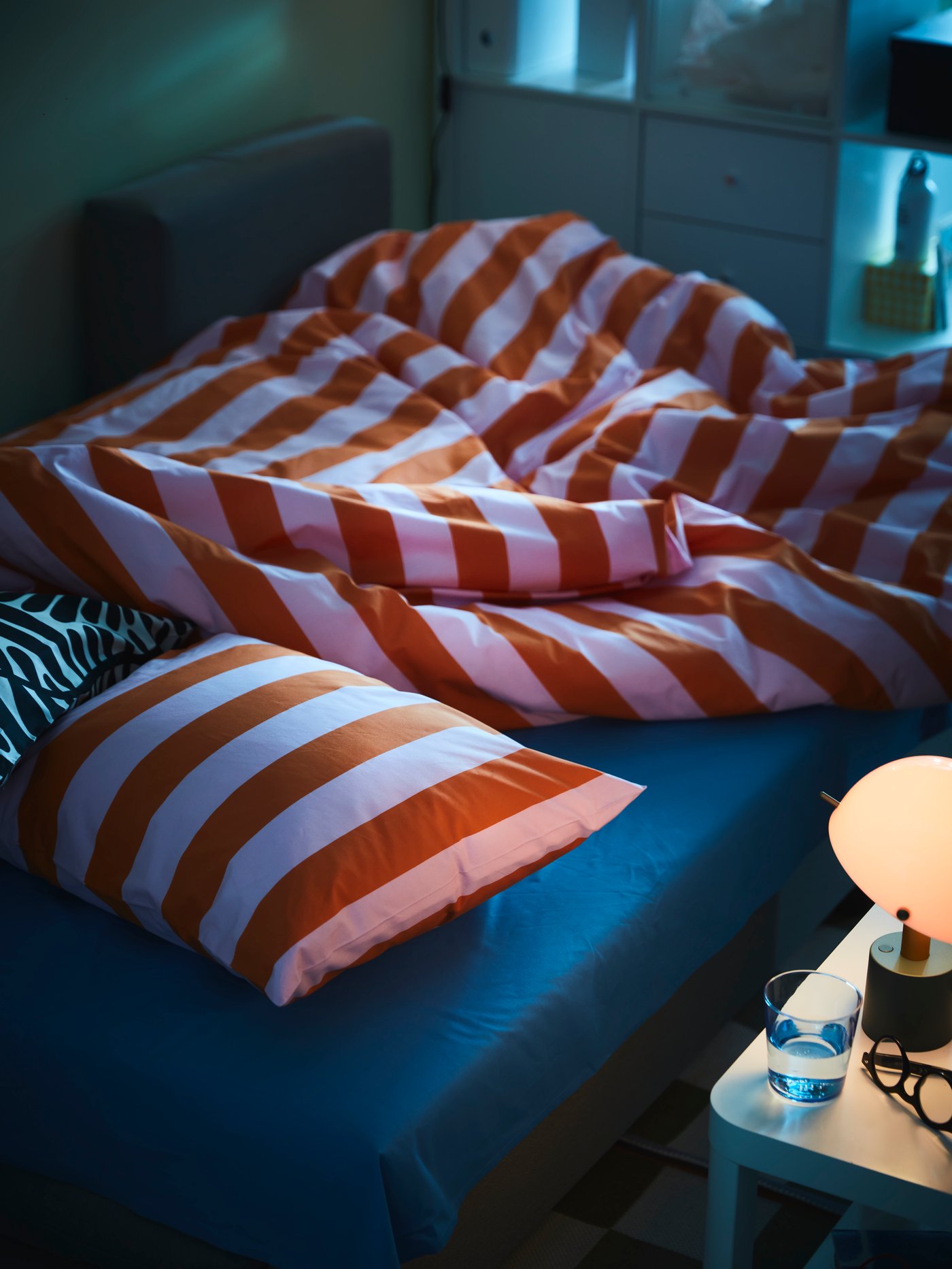 Obäddad säng med orange‑ och vitrandigt SLÅNHÖSTMAL påslakan och kudde i ett dunkelt sovrum. Ett nattduksbord med lampa och ett glas vatten står bredvid sängen.