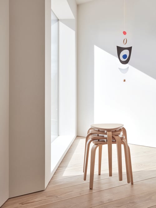 Une pièce blanche vide avec une grande fenêtre, une décoration suspendue et un ensemble de quatre tabourets en bouleau empilés les uns sur les autres.