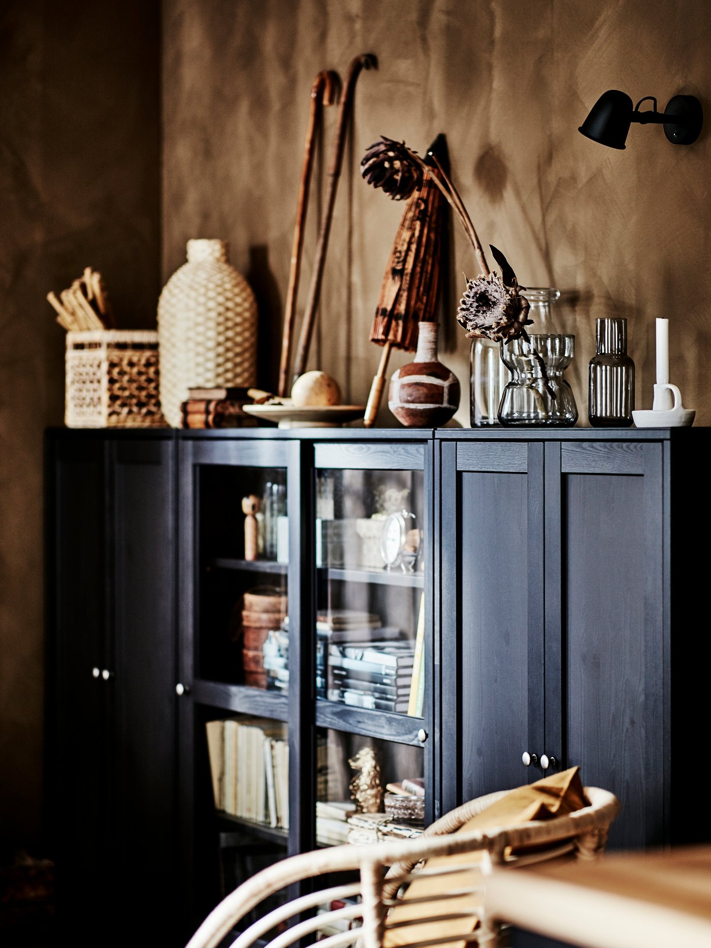 A wooden glass door cabinet containing various items, with diverse display ornaments on top, and a bamboo chair.