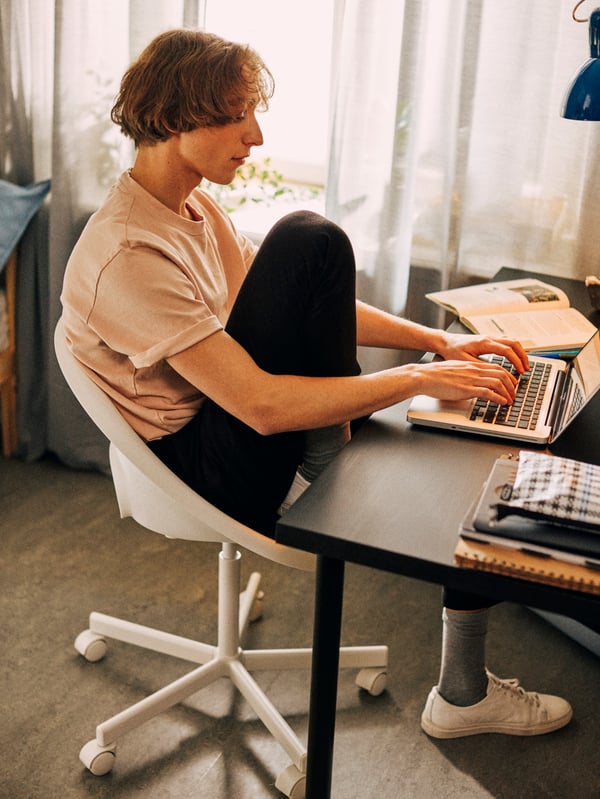 A boy sitting on a white LOBERGET/BLYSKÄR swivel chair and typing on a laptop placed on a LINNMON/ADILS table.