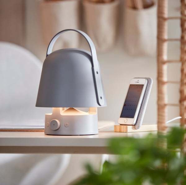 A VAPPEBY Bluetooth speaker lamp, shining its light, stands next to a mobile phone in a NORDMÄRKE wireless charging stand.