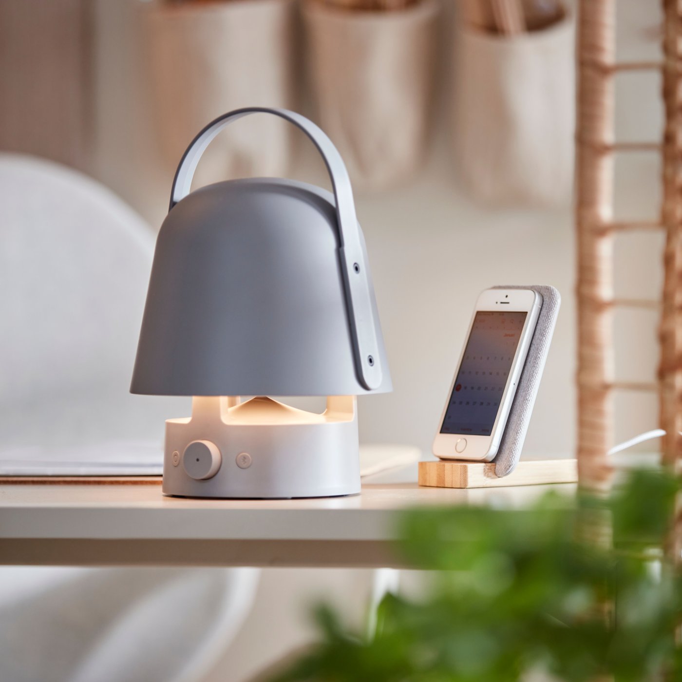 Eine eingeschaltete VAPPEBY Leuchte mit Bluetooth-Lautsprecher steht neben einem Mobiltelefon, das in einem kabellosen NORDMÄRKE Ladehalter geladen wird.