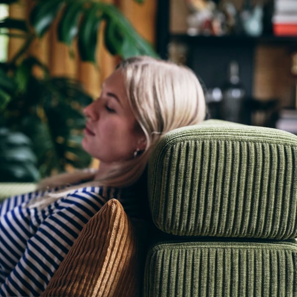 A woman peacefully reclining her head on a dark yellow-green JÄTTEBO headrest cushion placed on top of a JÄTTEBO sofa.