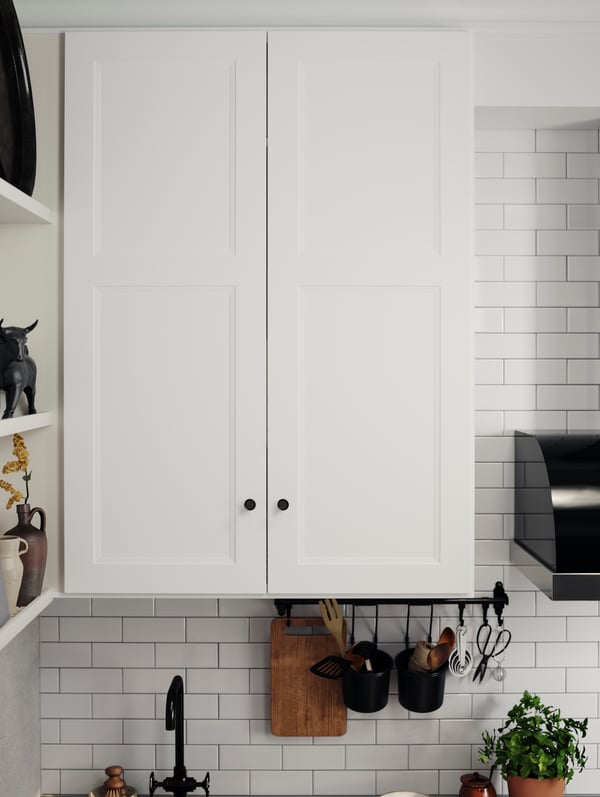 AXSTAD matte white kitchen cabinet doors with black knobs above a kitchen rail holding utensils and a cutting board.