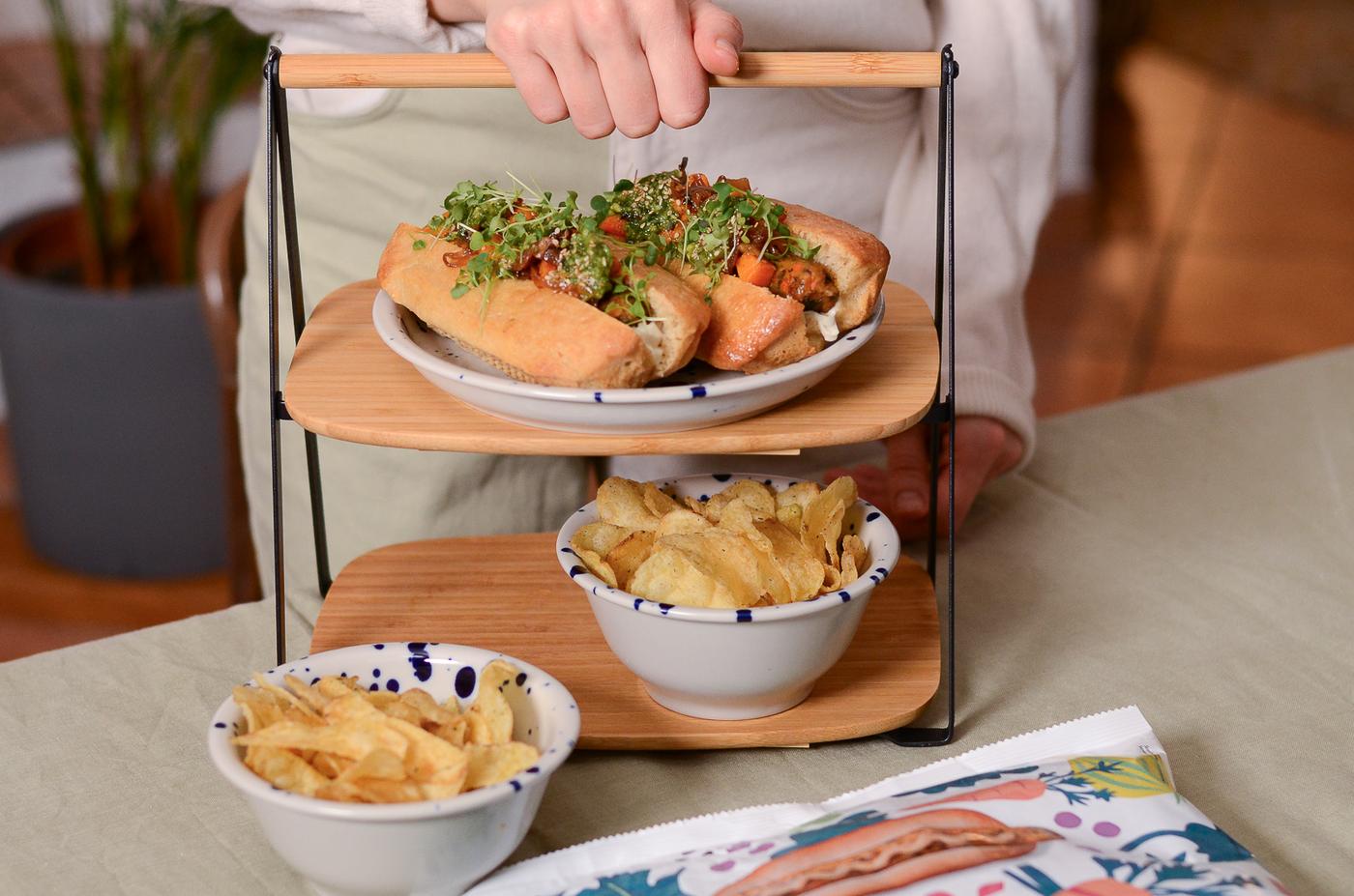 Una donna tiene in mano un vassoio con due hot dog e un contorno di patatine fritte, pronta a servire un pasto semplice.