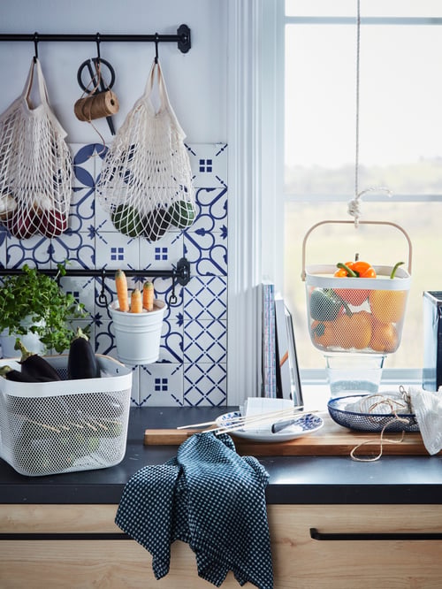 A RISATORP wire basket filled with aubergines on a kitchen countertop, with a beech chopping board in front of a window.