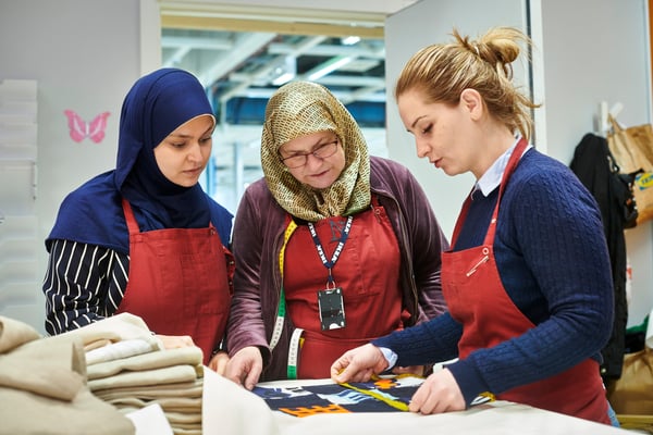 Three co-workers from Yalla Trappan sewing studio in Malmö, Sweden, measuring some fabric while wearing red aprons.