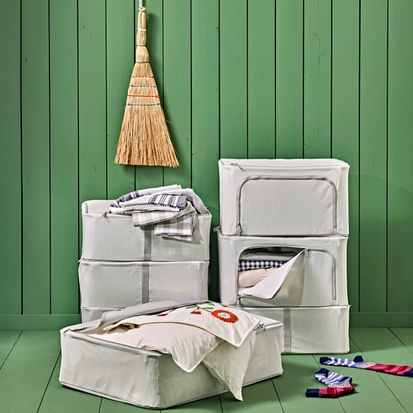 Several HEMMAFIXARE storage cases stacked against a green wall, one open on the floor showing home textiles inside.
