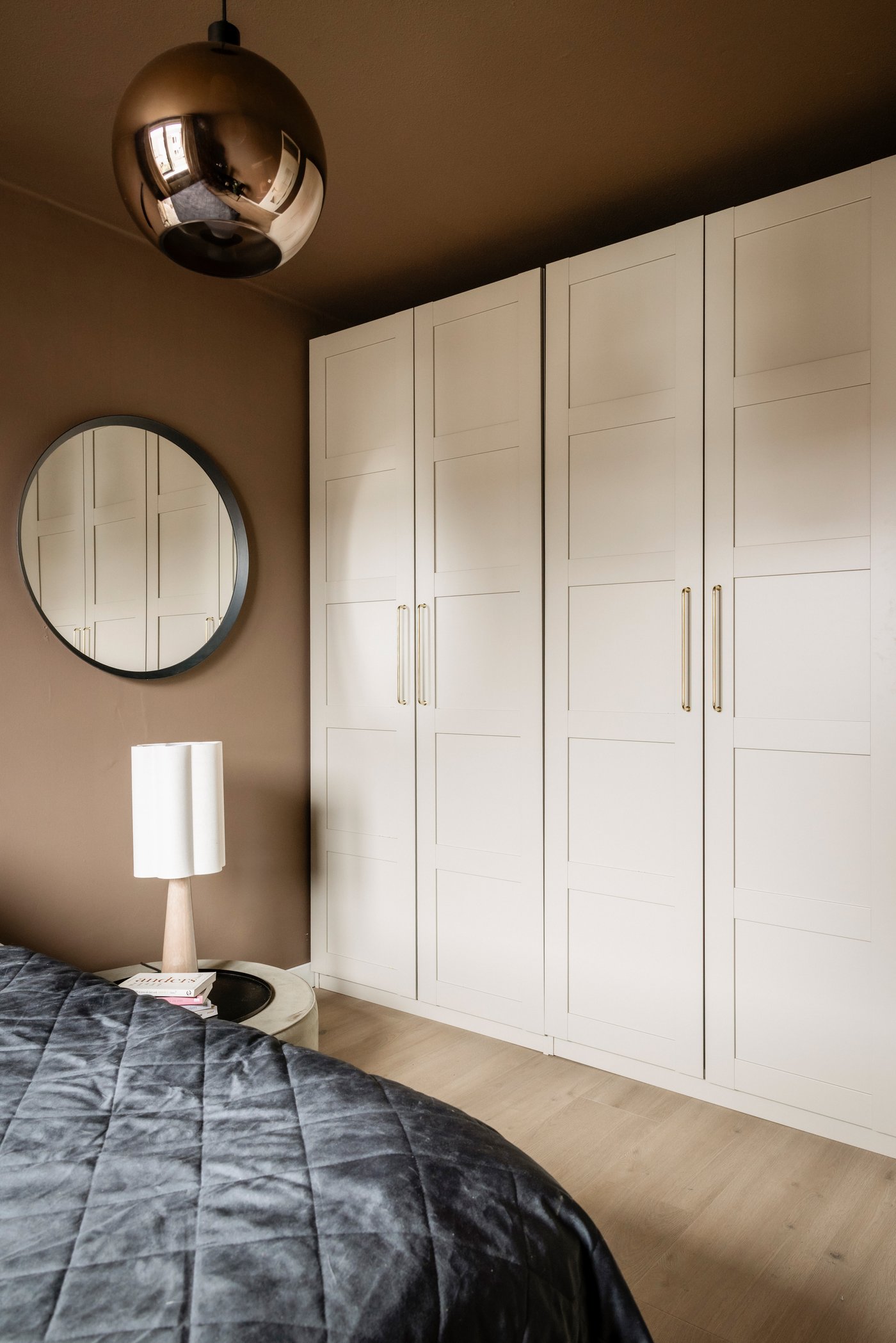 Wand van een slaapkamer waartegen een grote, beige PAX kledingkast staat, met gouden greepjes. Aan de muur hangt een ronde, zwarte spiegel.