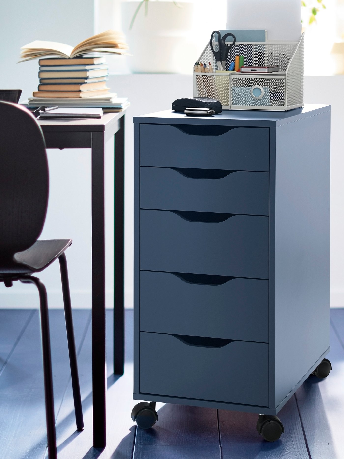 A black-blue ALEX drawer unit on castors next to a desk that has a pile of books on top.