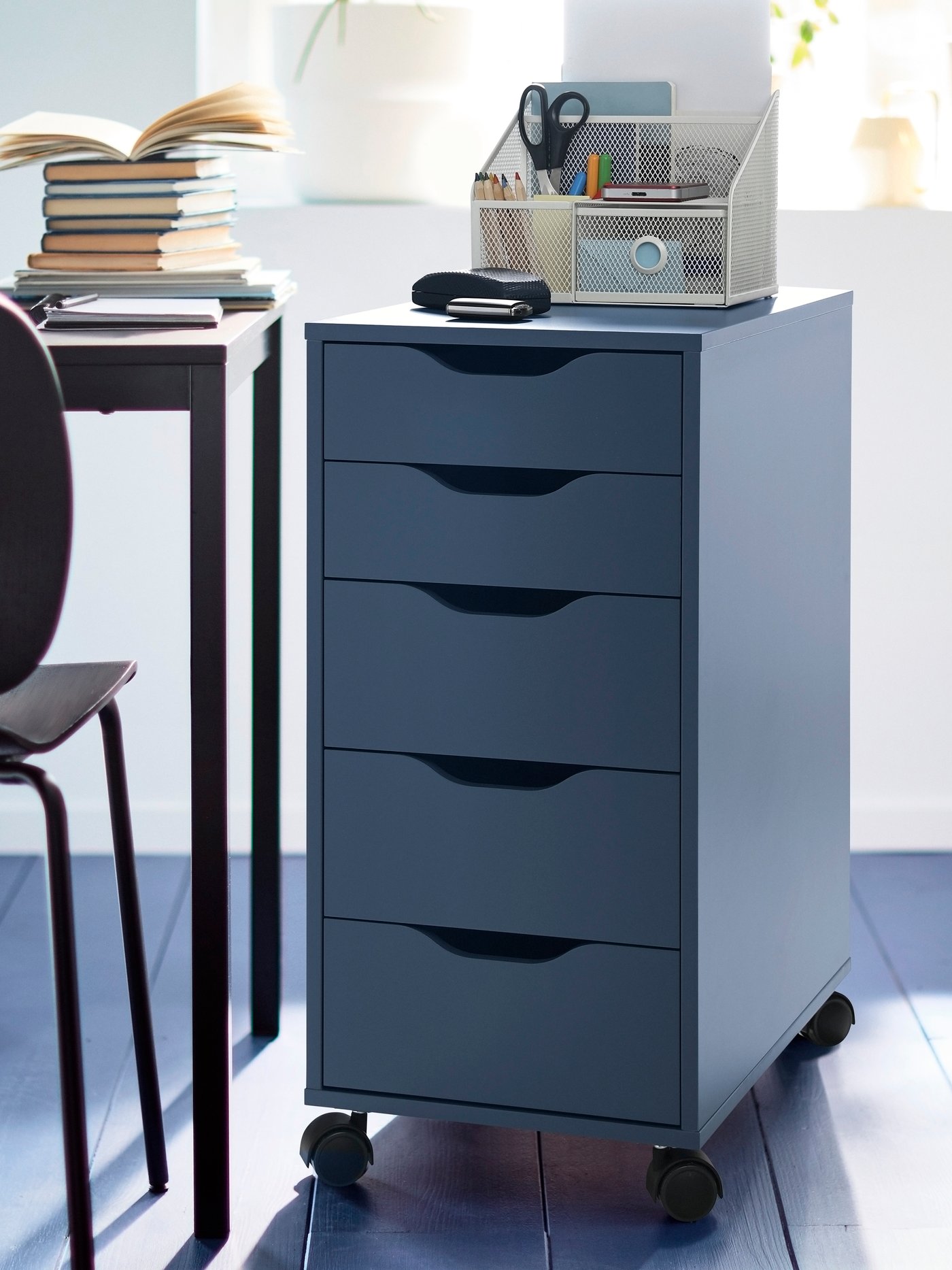 A black-blue ALEX drawer unit on castors next to a desk that has a pile of books on top.