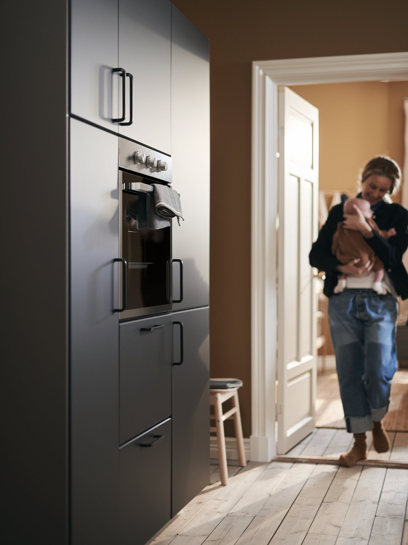 A mother cradles her baby as she enters the kitchen, approaching a wall of cabinets with KUNGSBACKA fronts.