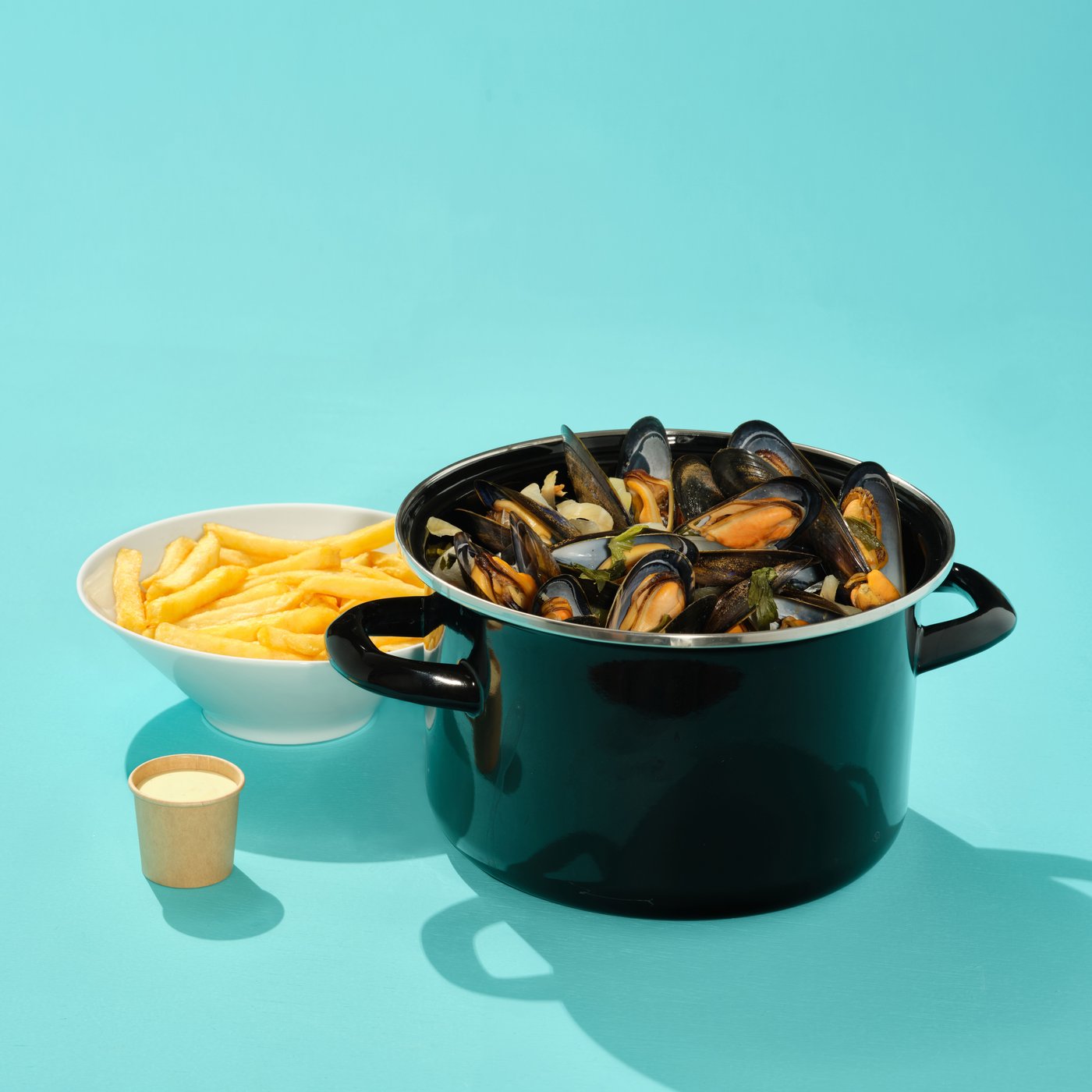 Mussels in a black pot with fries and sauce on a blue background, simple presentation.