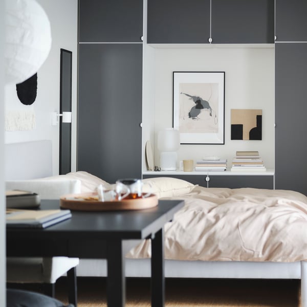 Uno specchio in una cornice LOMVIKEN nera. In fondo alla stanza una combinazioni di mobili grigio scuri con in mezzo un quadro di muro con dei quadri appessi e dei libri appoggiati al mobile. All'interno della stanza un letto con lenzuola rosa.
