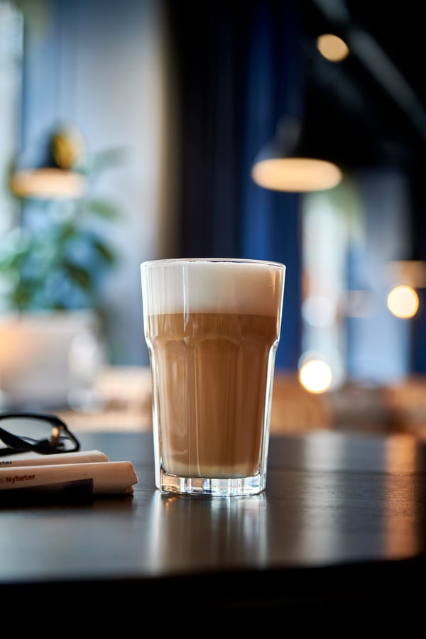 Kaffeglas med skum på mörkt bord, glasögon och penna bredvid, suddig bakgrund med lampor och växter.