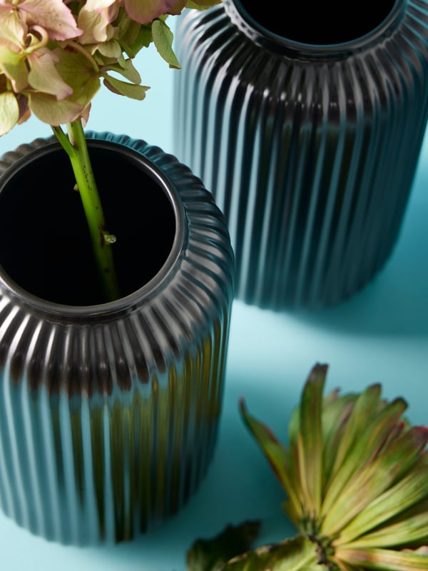 Two black STILREN vases, one holding a flower, against a light blue backdrop. There is a flower next to the vases.