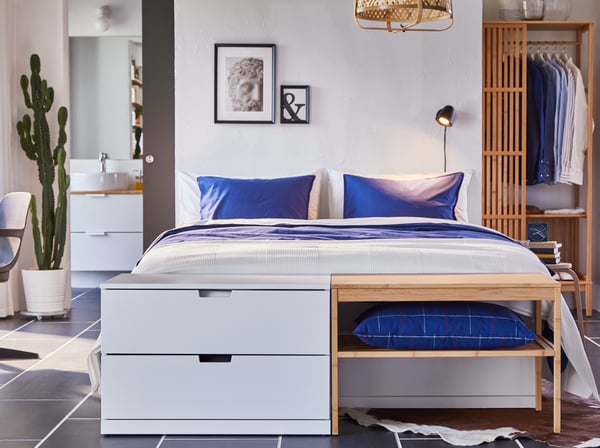 Quarto com uma estrutura de cama em branco, roupa de cama azul e um candeeiro suspenso, uma secretária, um roupeiro e prateleiras de bambu.