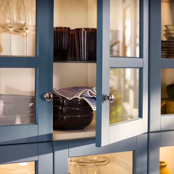A dining room cabinet with glass doors in LERHYTTAN blue has dining room items stored on shelves inside.