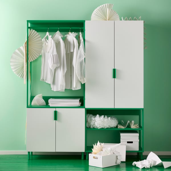 A white and green PLATSA wardrobe with 4 doors sits on a green floor against a green wall. It holds clothes, boxes, and more.