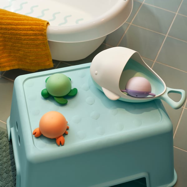 The various bath toys in the ÄNGSHUMLA collection on top of a light blue bathroom stool, beside a white baby bath.