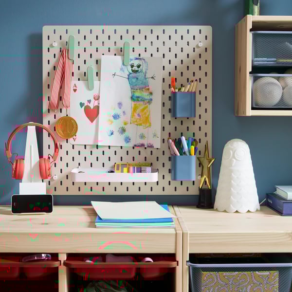 A beige SKÅDIS pegboard with colourful accessories holding pens and drawings placed on a blue wall above storage in pine.