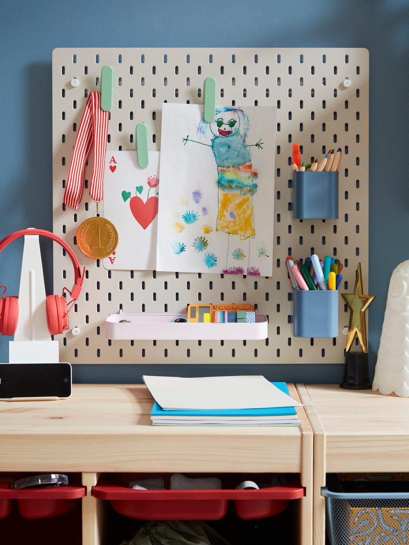 A beige SKÅDIS pegboard with colourful accessories holding pens and drawings placed on a blue wall above storage in pine.